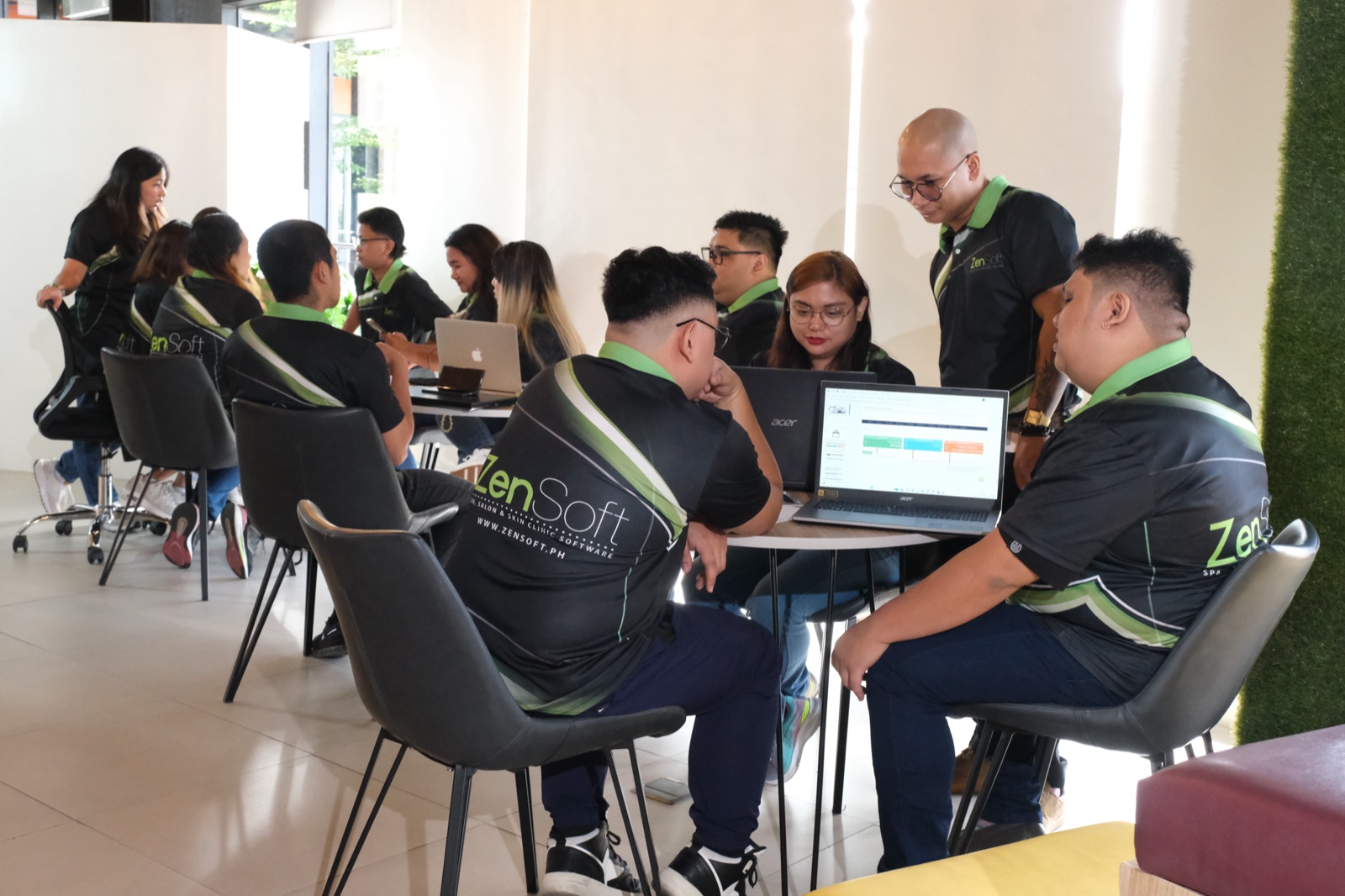 A ZenSoft team briefing in the lounge, staff seated at tables with their laptops